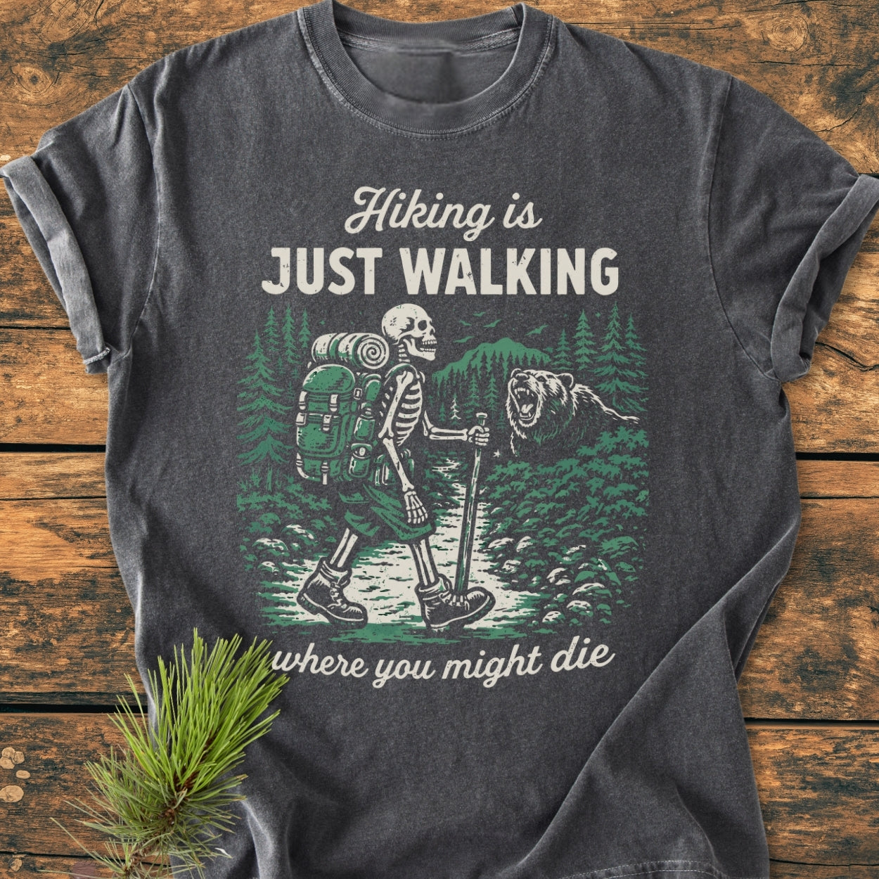 Hiking Is Just Walking - Vintage Wash Tee features a skeleton with a backpack hiking through a forest scene with a bear, printed on a soft, relaxed-fit dark-gray T-shirt.
