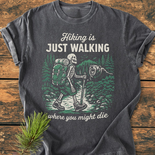 Hiking Is Just Walking - Vintage Wash Tee features a skeleton with a backpack hiking through a forest scene with a bear, printed on a soft, relaxed-fit dark-gray T-shirt.