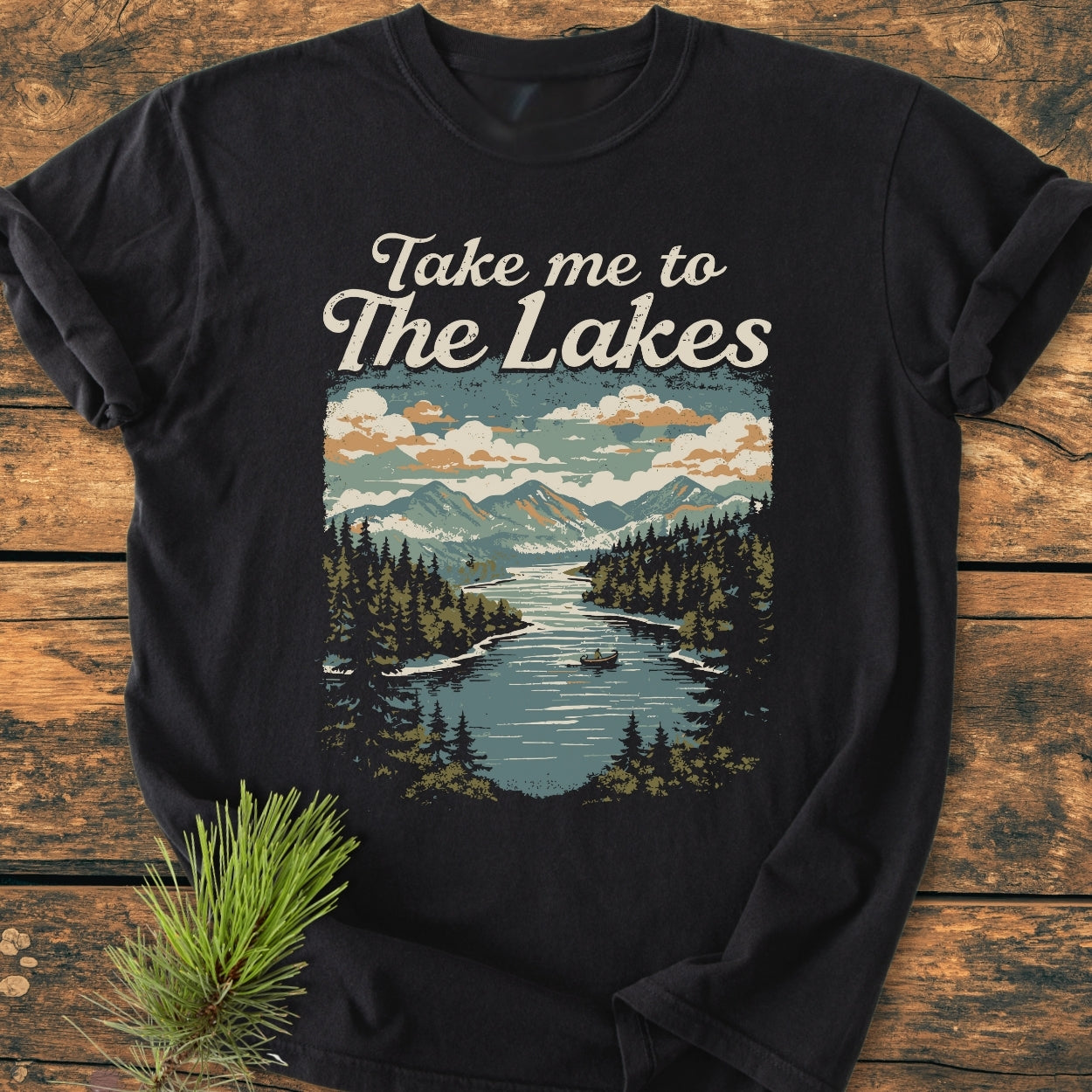 Take Me To The Lakes - Vintage Wash Tee featuring a mountain lake illustration, laid on wood. Soft, relaxed fit, 100% ring-spun cotton, emphasizing durability and comfort with double-needle stitching.