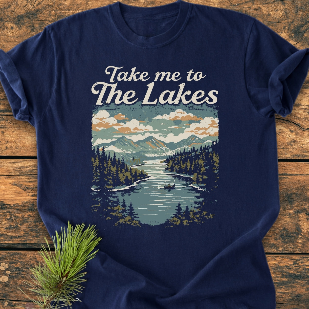 Take Me To The Lakes - Vintage Wash Tee features a mountain lake graphic with snow-capped peaks and a rowboat, accentuating its outdoorsy charm, laid on a wooden surface with a pine sprig.