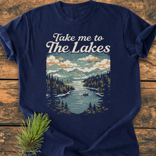 Take Me To The Lakes - Vintage Wash Tee features a mountain lake graphic with snow-capped peaks and a rowboat, accentuating its outdoorsy charm, laid on a wooden surface with a pine sprig.