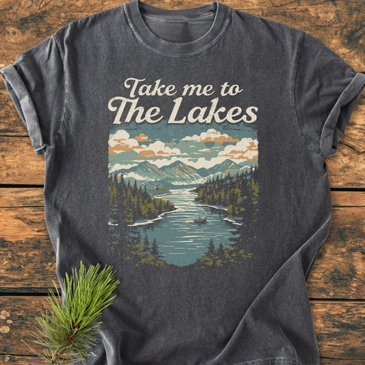 Take Me To The Lakes - Vintage Wash Tee: A dark-charcoal T-shirt featuring a vintage-style mountain lake print with a canoeist, laid flat on a wooden surface with a pine sprig beside it.