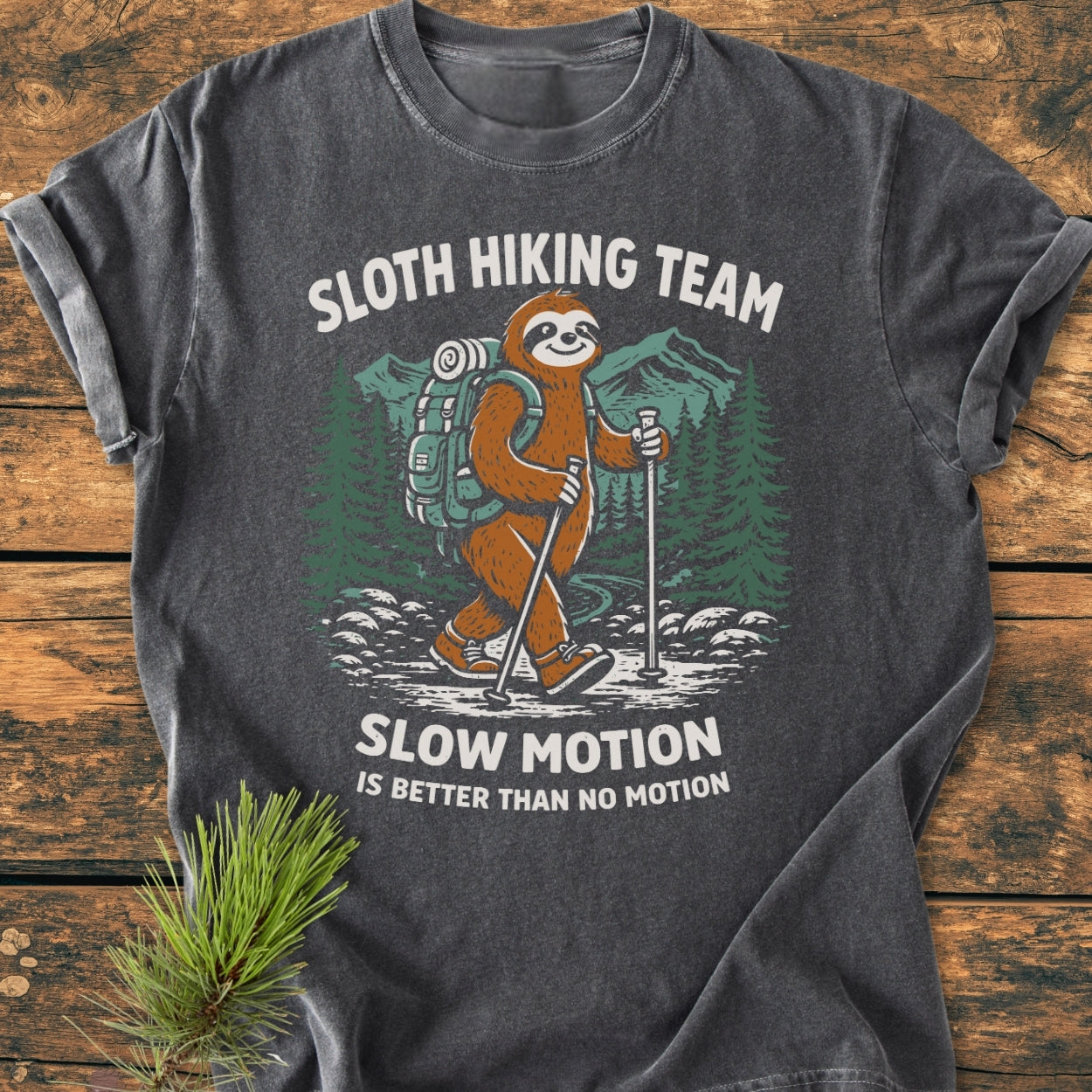 Sloth Hiking Team - Vintage Wash Tee features a cartoon sloth in hiking gear with trekking poles, set against pine trees and mountains, captioned with playful text. Laid on a wooden surface with a pine branch.
