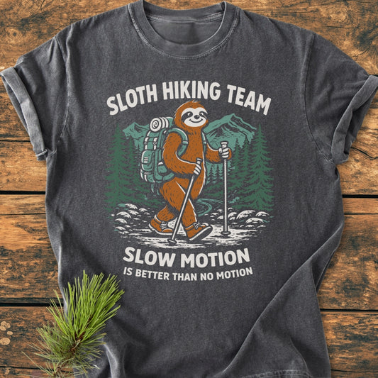 Sloth Hiking Team - Vintage Wash Tee features a cartoon sloth in hiking gear with trekking poles, set against pine trees and mountains, captioned with playful text. Laid on a wooden surface with a pine branch.