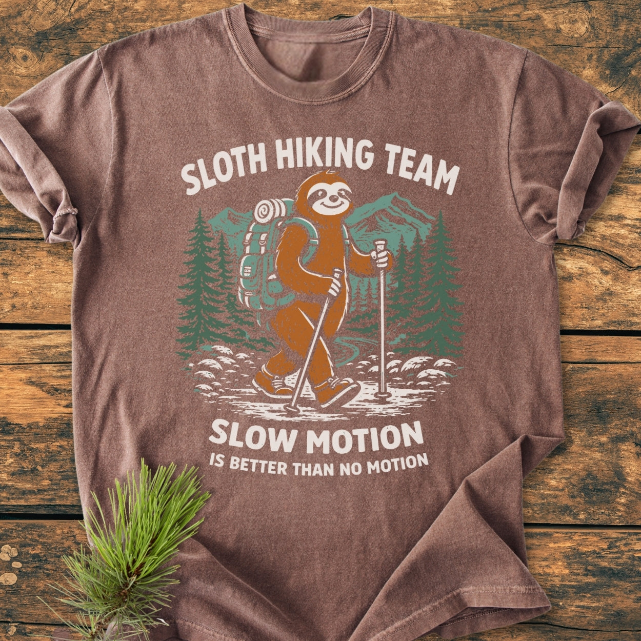 Sloth Hiking Team - Vintage Wash Tee featuring a cartoon sloth in hiking gear with the phrase Slow Motion Is Better Than No Motion, laid on a wooden surface, showcasing its relaxed fit and durability.
