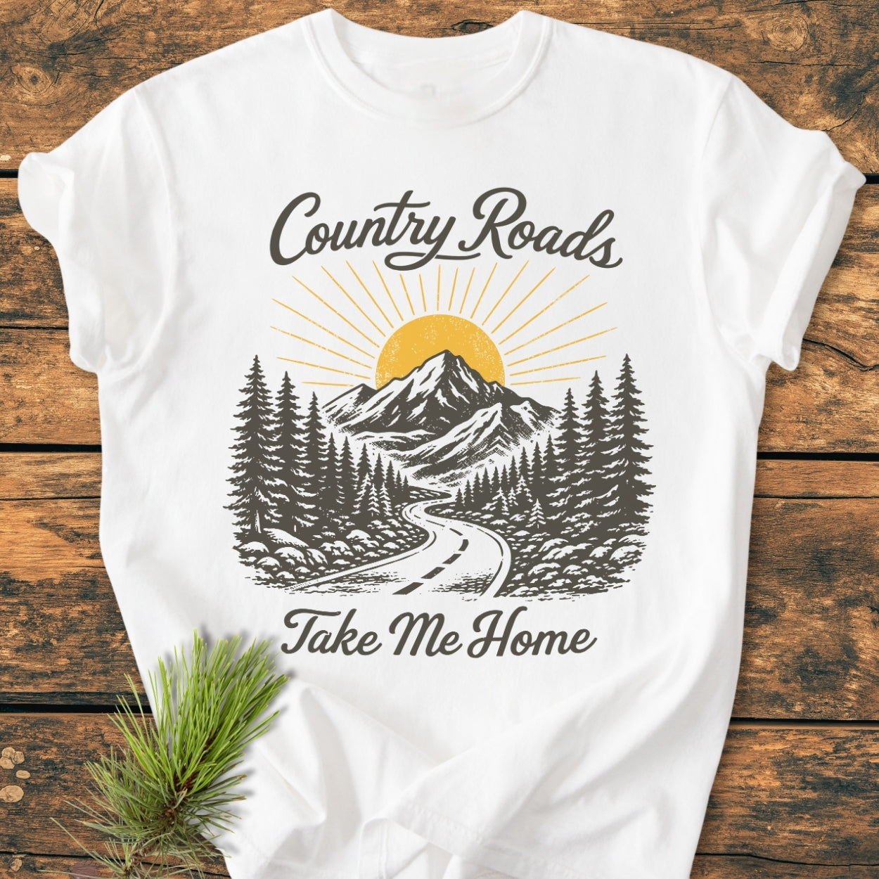 Country Roads - Vintage Wash Tee features a stylized mountain-and-forest scene with a winding road on a soft, white tee, accented by a pine branch. Text reads “Country Roads” and “Take Me Home.”