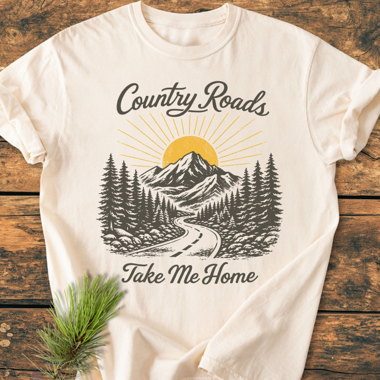 Country Roads - Vintage Wash Tee featuring a nature scene with a sun, mountains, pine forest, and winding road, with “Country Roads” and “Take Me Home” in cursive on a wooden surface.