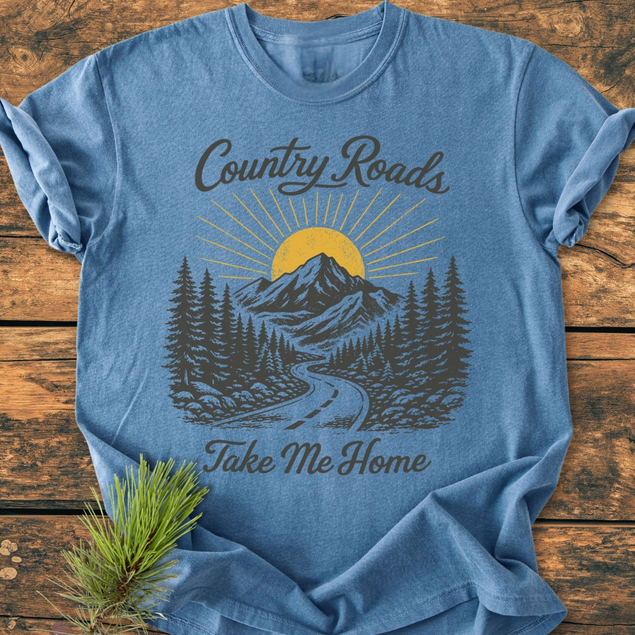 Country Roads - Vintage Wash Tee: Heather-blue T-shirt featuring a scenic landscape with a winding road and mountains. Text reads “Country Roads, Take Me Home.” Pine sprig accentuates the natural theme.