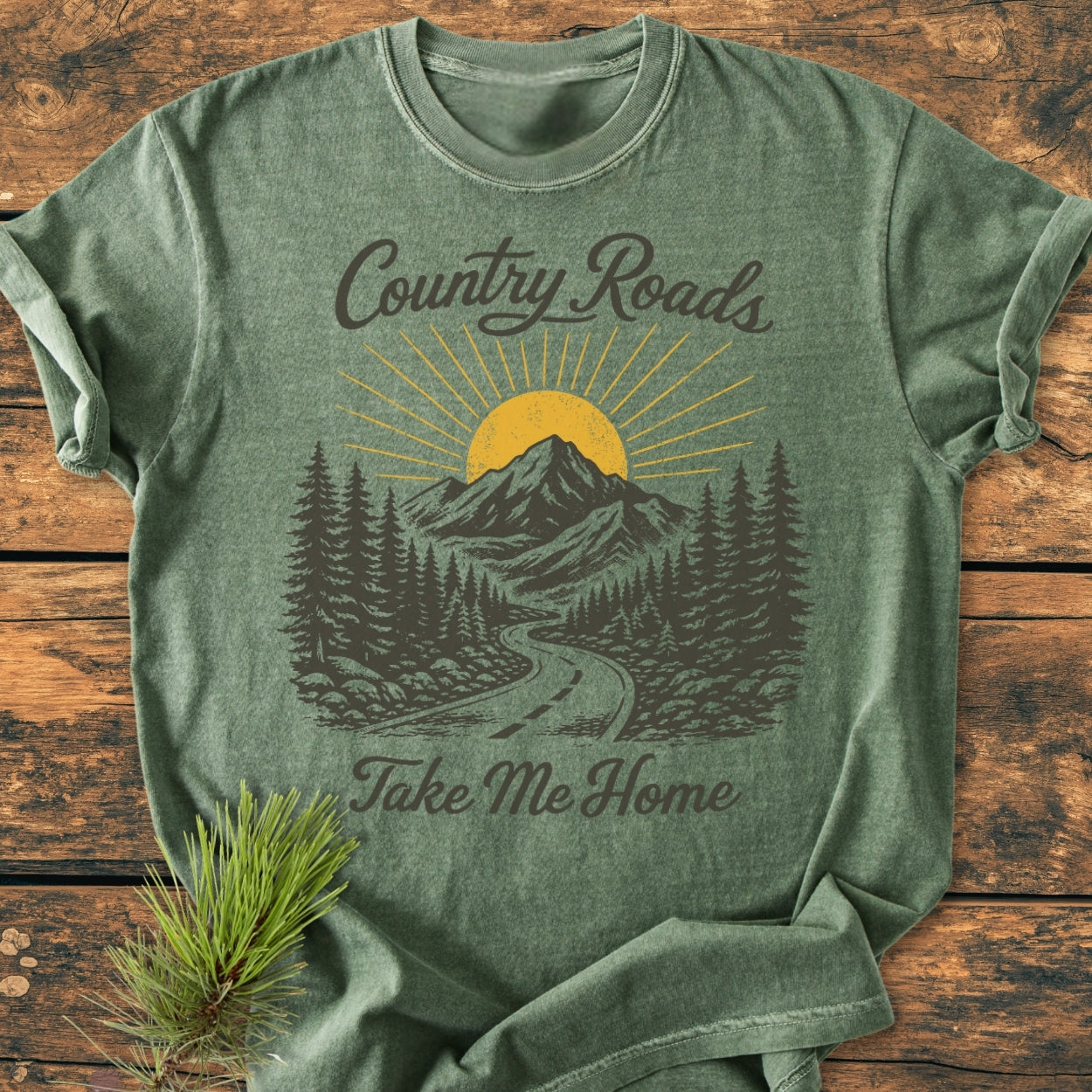 Country Roads - Vintage Wash Tee featuring a scenic graphic of a country road, pine trees, and mountain with sun, laid on wood. Includes text “Country Roads” and “Take Me Home,” with pine branch accent.