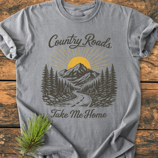 Country Roads - Vintage Wash Tee, heather-gray, features a scenic print with a road, pine trees, mountains, and sun. Laid flat on wood, it includes “Country Roads, Take Me Home” text.