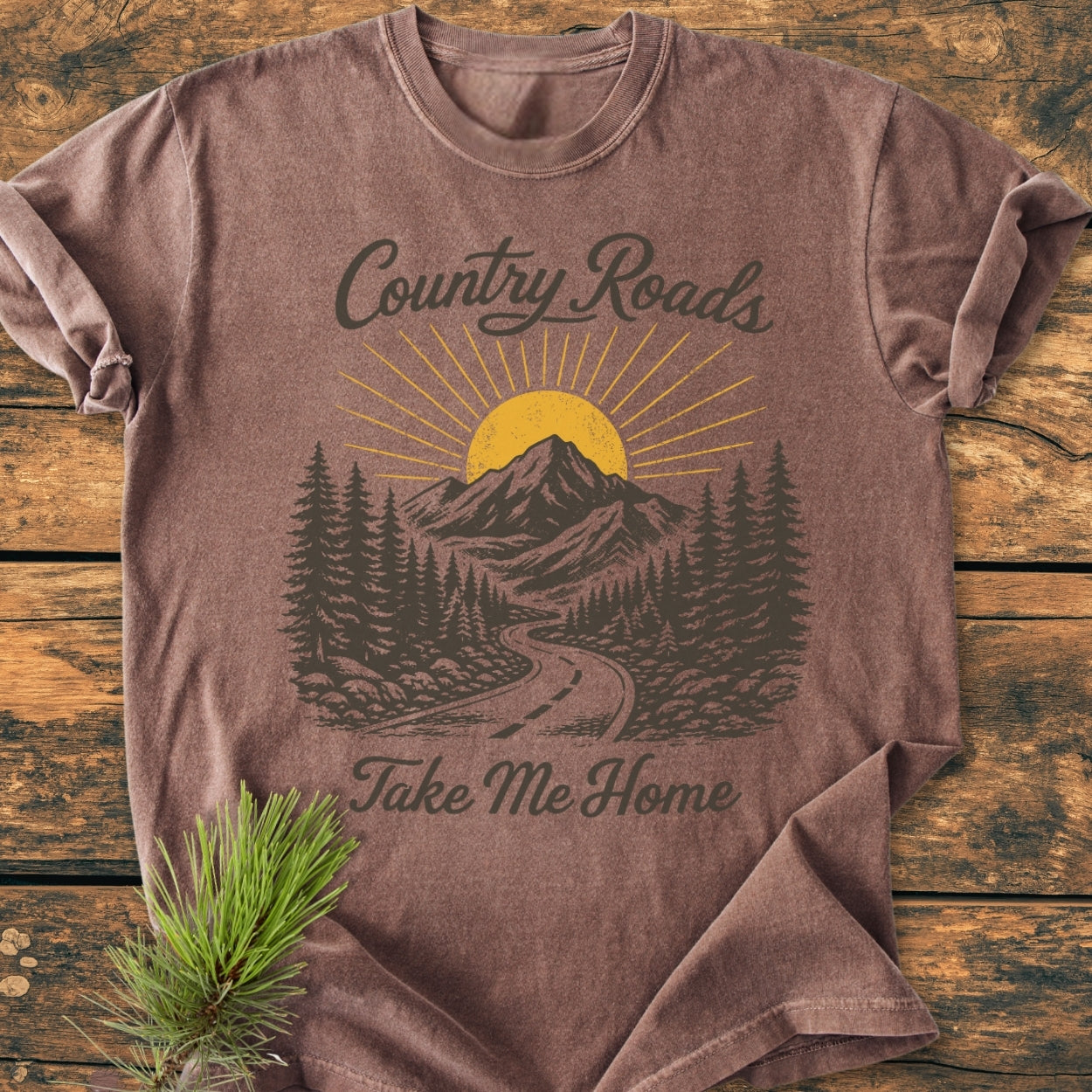 Country Roads - Vintage Wash Tee featuring a rustic graphic of a country road, pine trees, and mountains under a rising sun, with Country Roads and Take Me Home script, complemented by pine sprigs.