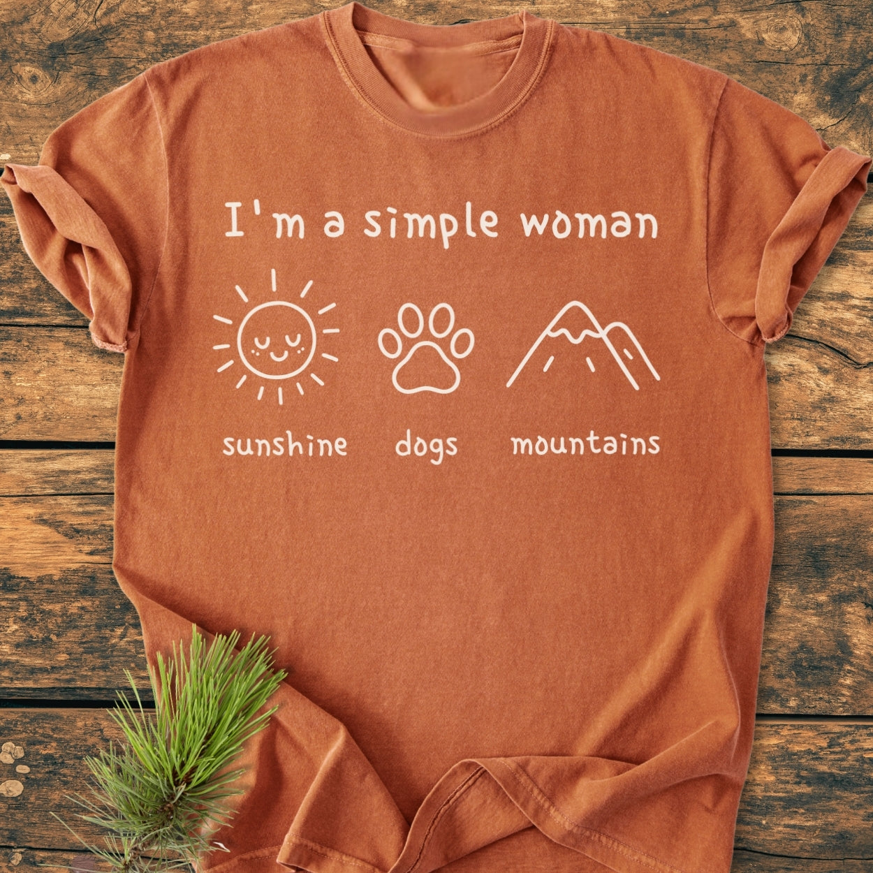 I'm A Simple Woman - Sunshine, Dogs, Mountains - Vintage Wash Tee: Rust-orange crew-neck T-shirt with white lettering and icons on chest, styled with a pine sprig on a wooden surface.