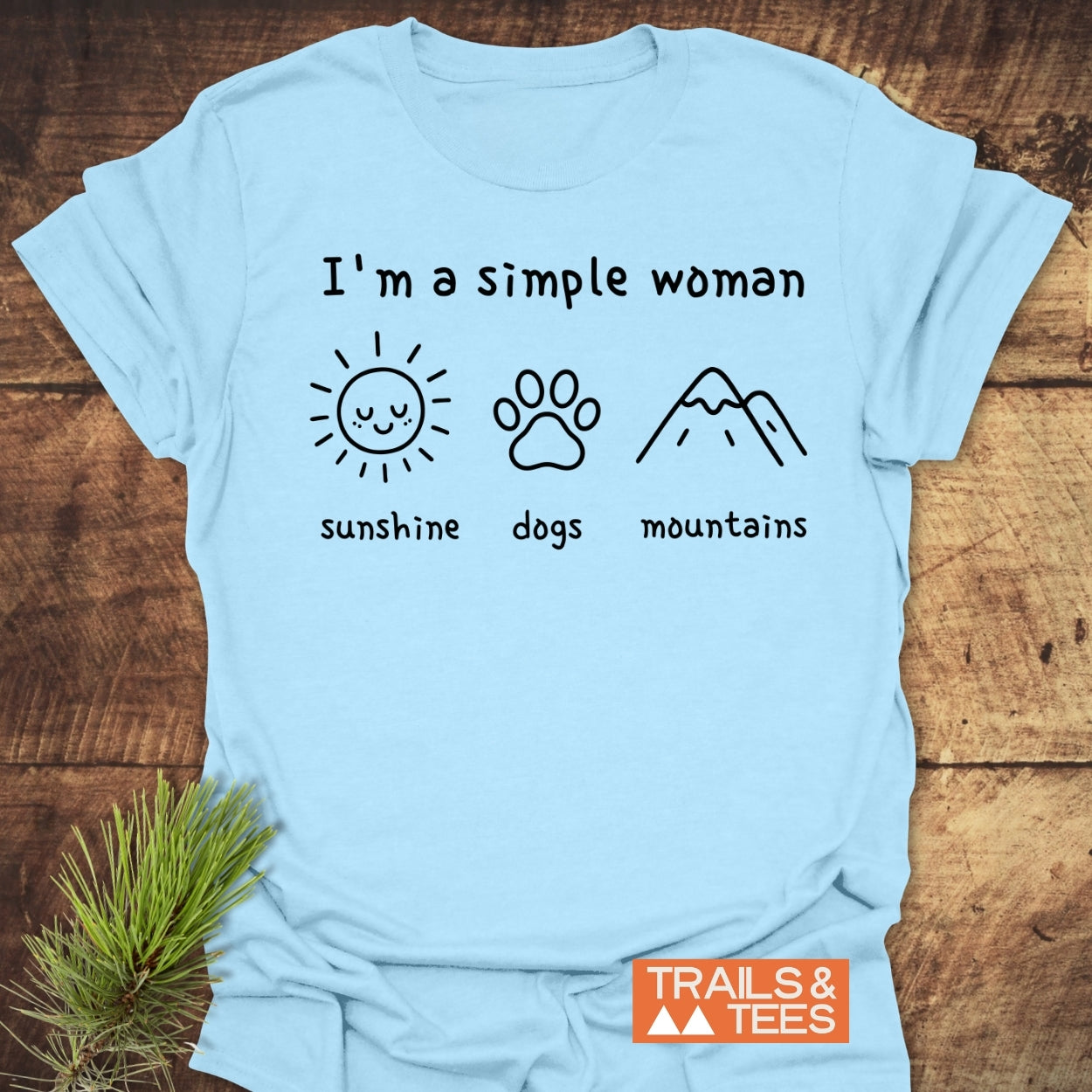 I'm A Simple Woman - Sunshine, Dogs, Mountains T-Shirt featuring line-drawn icons of a sun, paw print, and mountain, showcasing Trails & Tees' comfort-focused, outdoor-inspired design on a wooden surface.