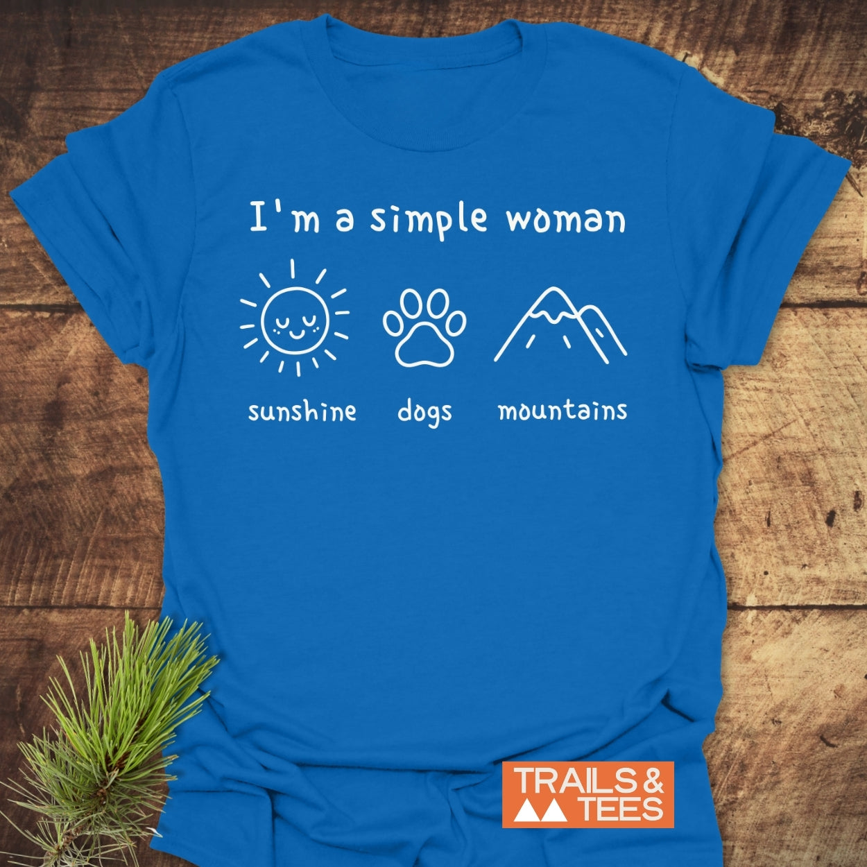 I'm A Simple Woman - Sunshine, Dogs, Mountains T-Shirt featuring a bright blue design with three icons and a TRAILS & TEES logo, laid on wood with pine sprig beside it.