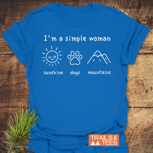 I'm A Simple Woman - Sunshine, Dogs, Mountains T-Shirt featuring a bright blue design with three icons and a TRAILS & TEES logo, laid on wood with pine sprig beside it.