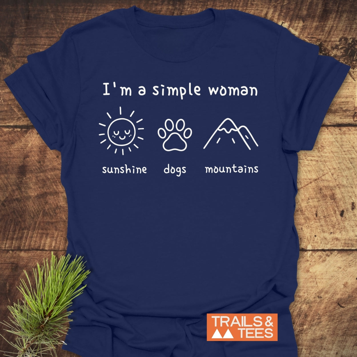 The I'm A Simple Woman - Sunshine, Dogs, Mountains T-Shirt features a white hand-drawn design on navy, highlighting sunshine, dogs, and mountains, with the Trails & Tees brand logo at the corner.