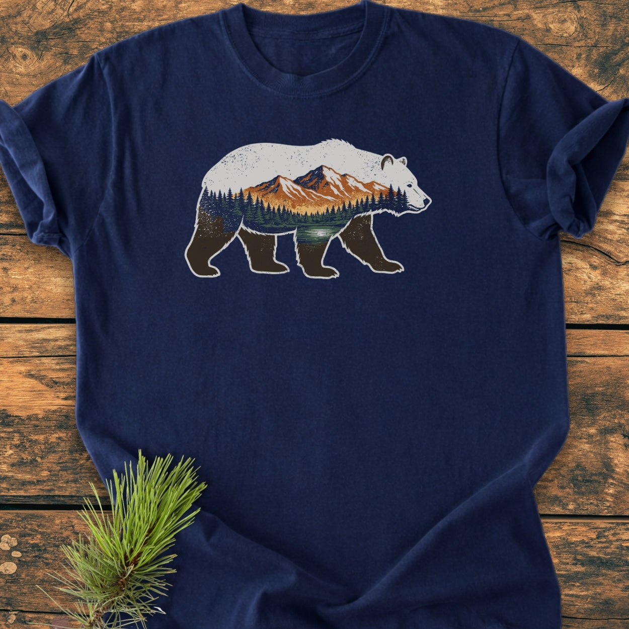 Mountain Within - Vintage Wash Tee: A navy T-shirt with a bear graphic depicting trees, a lake, and mountains. Laid on wood, it includes a sprig of evergreen, echoing wilderness themes.