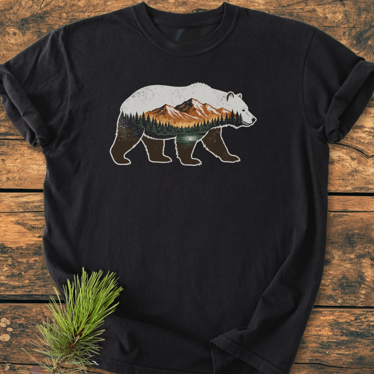 Mountain Within - Vintage Wash Tee features a walking bear graphic with a pine-forest scene on a black crew-neck t-shirt, laid out on a wooden surface with pine needles.