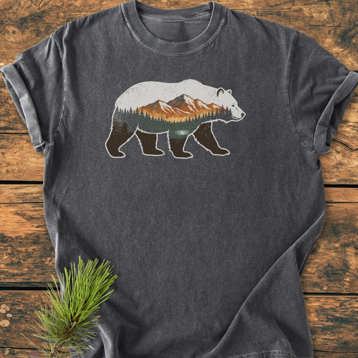 Mountain Within - Vintage Wash Tee features a bear silhouette with a forested mountain scene. Displayed on a wooden surface, this charcoal-gray t-shirt embodies comfort and style with its relaxed fit and durable stitching.