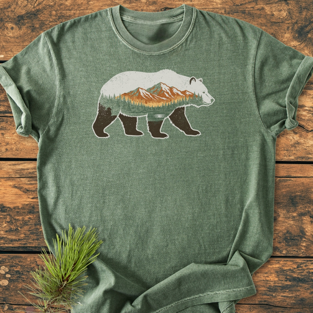 Mountain Within - Vintage Wash Tee featuring a bear silhouette with a pine and mountain landscape, laid flat on a wood-grain background, emphasizing its relaxed fit and durable, soft-washed fabric.