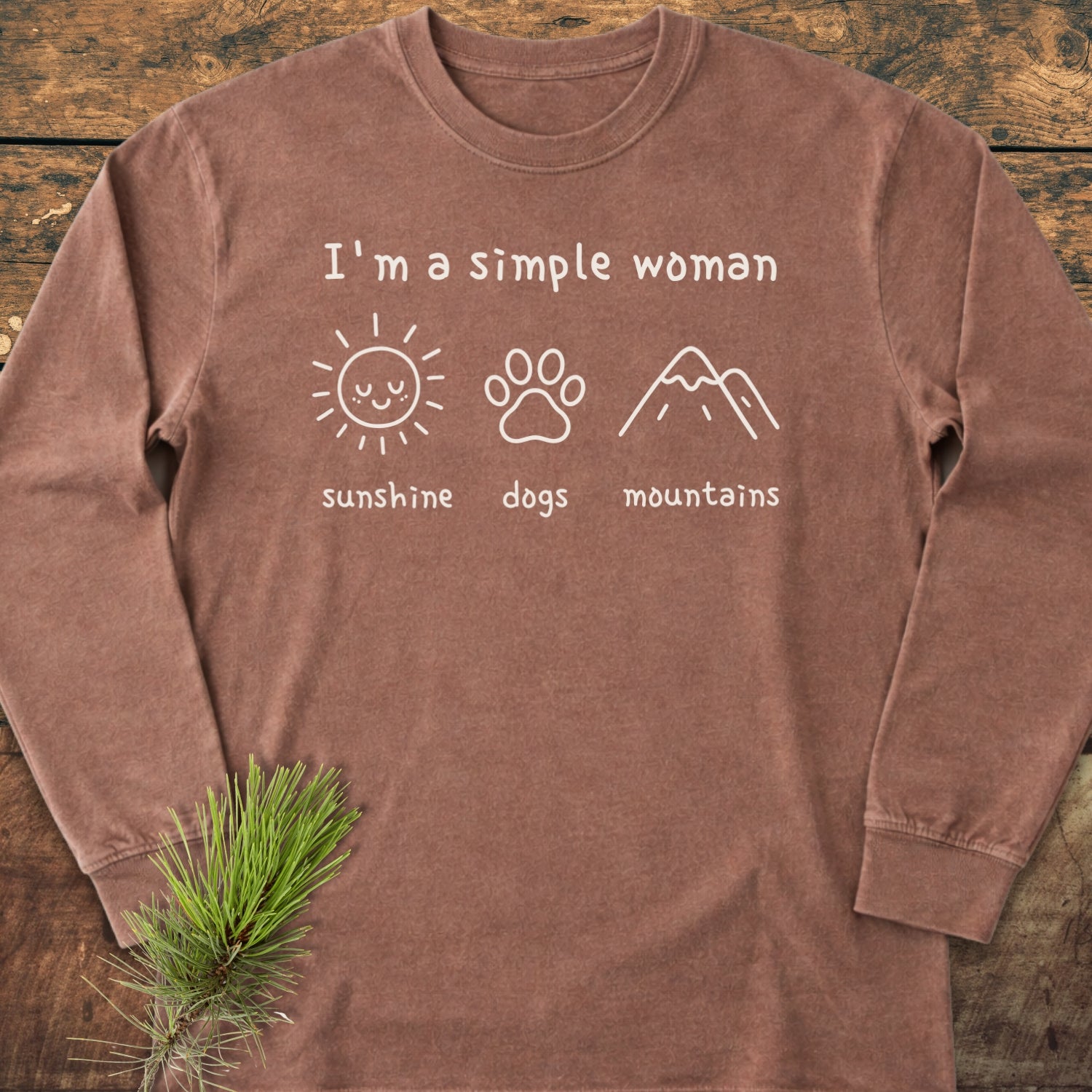 I'm A Simple Woman - Sunshine, Dogs, Mountains Long-Sleeve Tee featuring white text and icons on a brown cotton shirt, laid flat with a pine sprig accent, highlighting casual comfort and style.