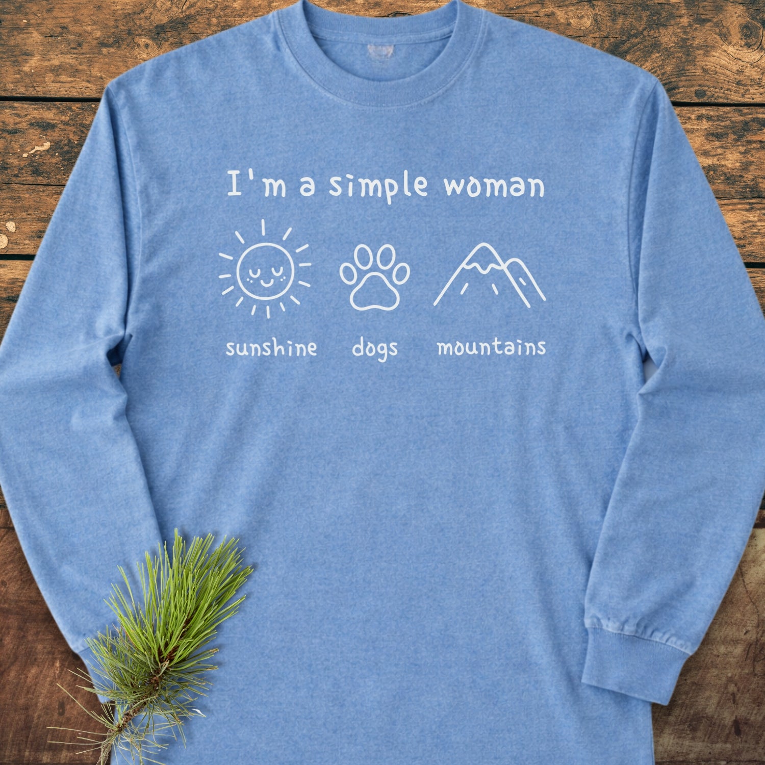 The I'm A Simple Woman - Sunshine, Dogs, Mountains Long-Sleeve Tee features white icons of a sun, dog paw, and mountains on a heather-blue shirt, styled with a pine sprig.