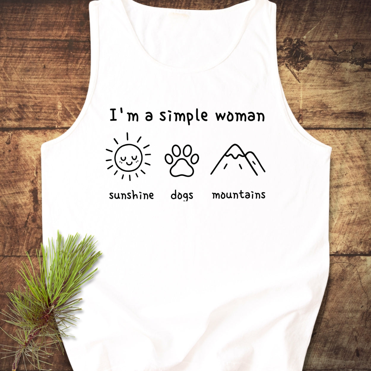 I'm A Simple Woman - Sunshine, Dogs, Mountains Tank Top features a white, sleeveless design with black line art and icons on a wooden surface, styled with a pine branch.