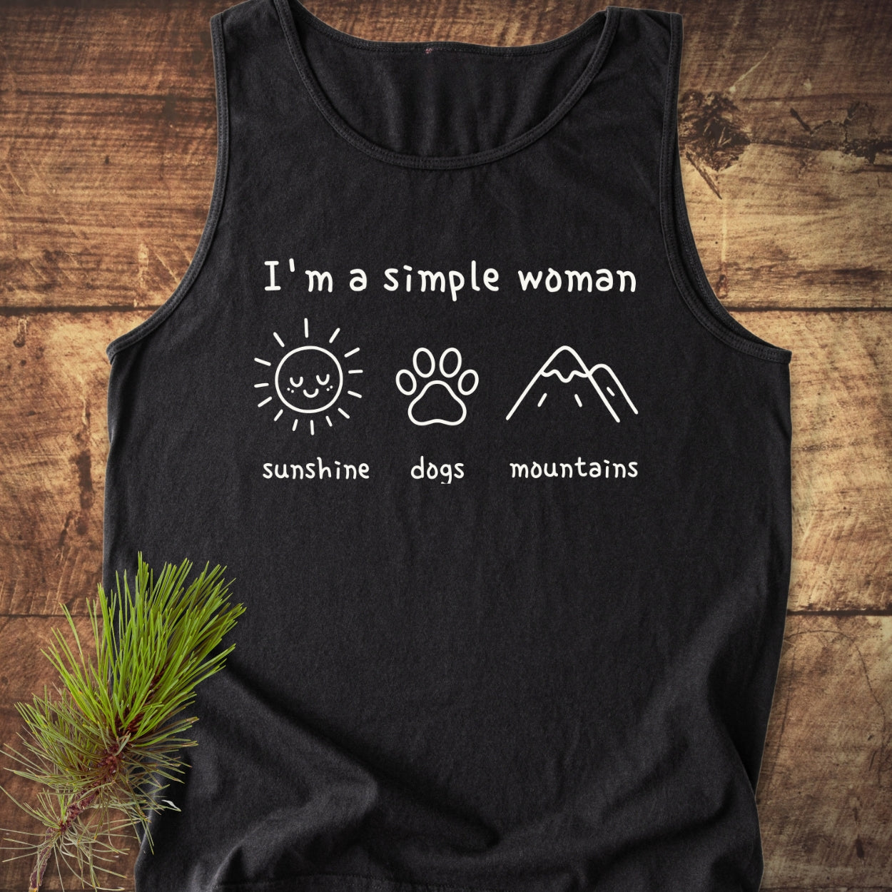 I'm A Simple Woman - Sunshine, Dogs, Mountains Tank Top featuring white graphics on a black tank, laid flat on wood with a pine branch. Comfortable, sustainable, 100% ring-spun US cotton, relaxed fit.