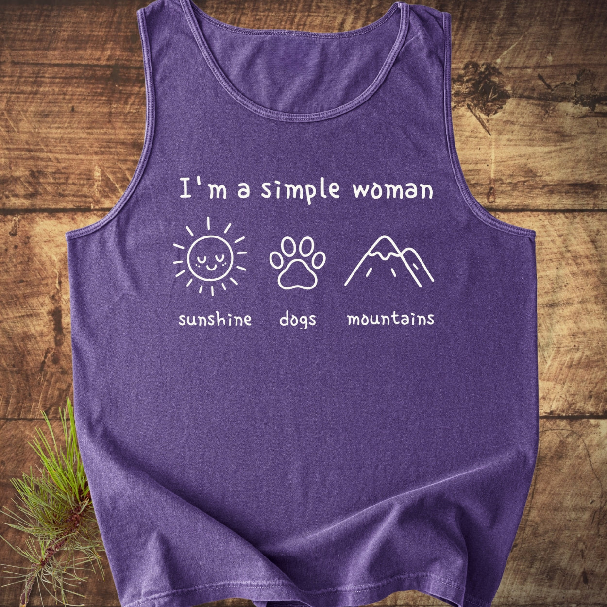 I'm A Simple Woman - Sunshine, Dogs, Mountains Tank Top on a wooden surface, featuring white lettering and icons of a sun, paw print, and mountains, with a pine sprig beside.