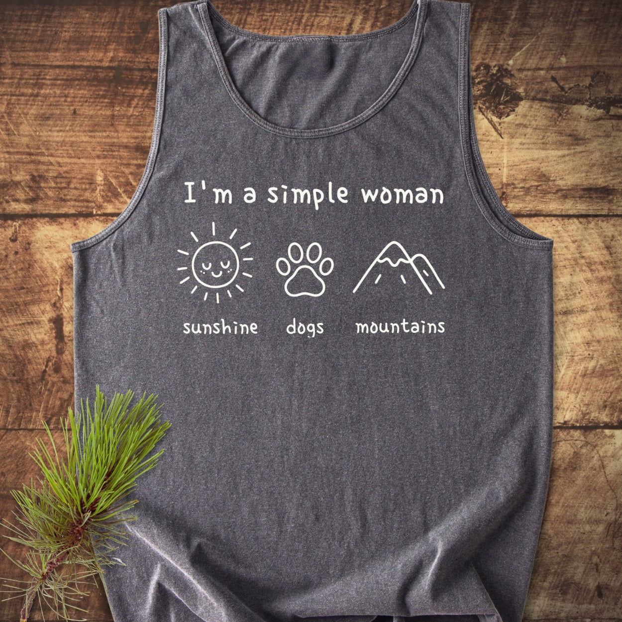 I'm A Simple Woman - Sunshine, Dogs, Mountains Tank Top: Heather-grey tank top with icons of a sun, dog paw, and mountains, labeled accordingly, laid on wood with a pine sprig beside it.