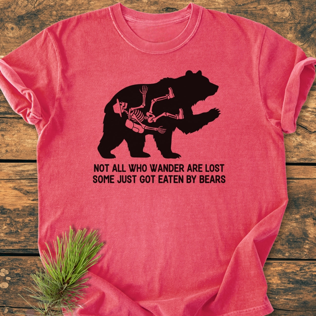 Not All Who Wander Are Lost - Vintage Wash Tee featuring a black bear silhouette with a hiker's skeleton on a pink-red background, laid on wood with pine sprigs, captioned humorously about wandering and bears.