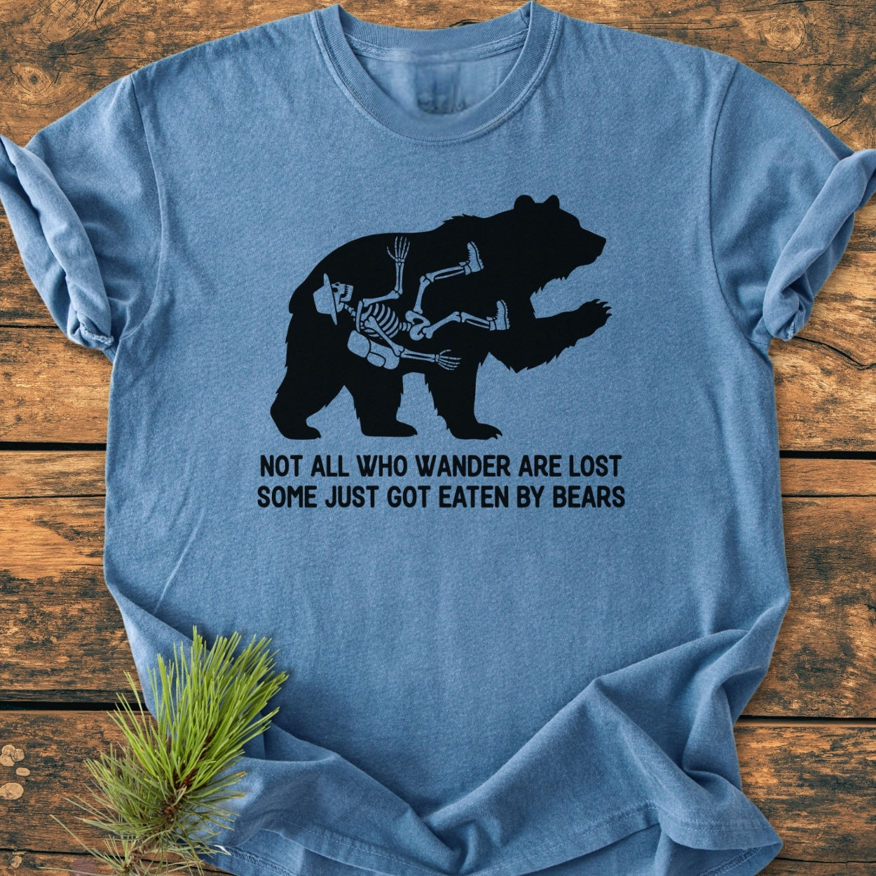 Not All Who Wander Are Lost - Vintage Wash Tee features a bear silhouette with a hiker's skeleton, printed on a laid-out light-blue T-shirt, emphasizing comfort and durability with a whimsical design.