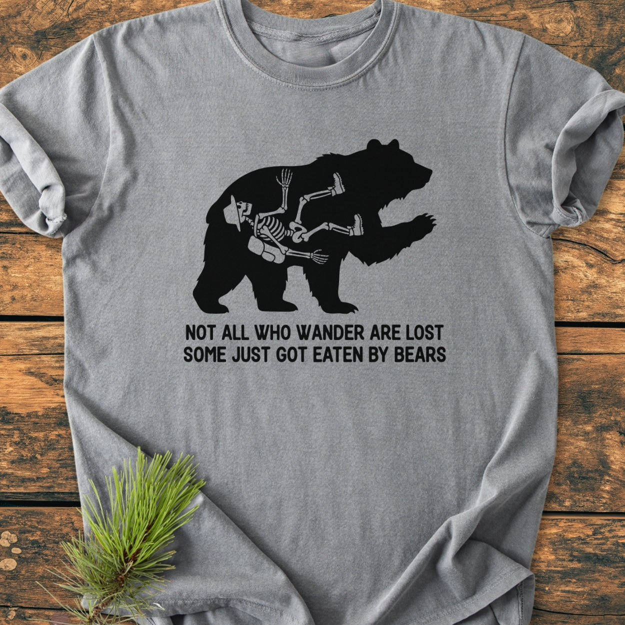 Heather-gray Not All Who Wander Are Lost - Vintage Wash Tee featuring a black bear silhouette with a hiker's skeleton inside, laid on wood with pine sprig. Caption reads humorous bear-themed hiking message.