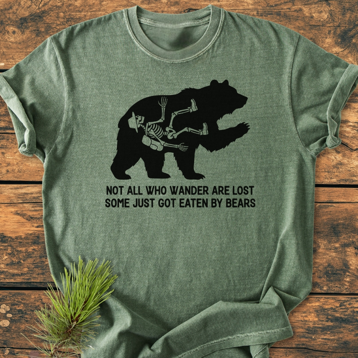 Not All Who Wander Are Lost - Vintage Wash Tee features a green T-shirt with a bear silhouette and hiker skeleton graphic, humorous caption, and pine sprig accent on a wooden surface.