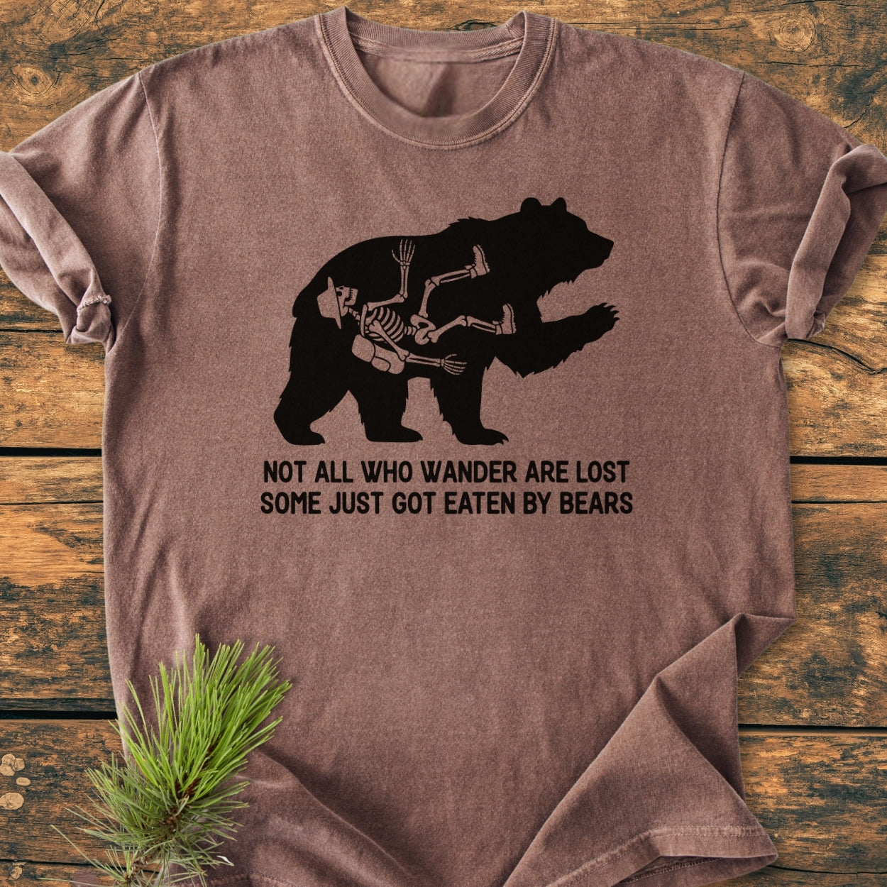 Not All Who Wander Are Lost - Vintage Wash Tee features a brown, crew-neck design with a bear silhouette and hiker's skeleton graphic, laid flat on a wooden surface with pine sprig accent.