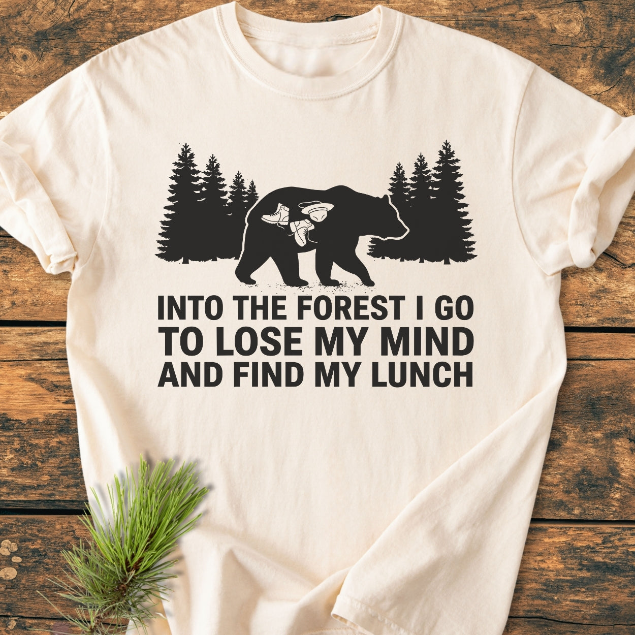 Into The Forest Bear - Vintage Wash Tee, featuring a black bear silhouette with pine trees, hiking boots, and a cooking pot graphic, slogan beneath. Laid out on a wooden surface, highlighting its soft, relaxed fit.
