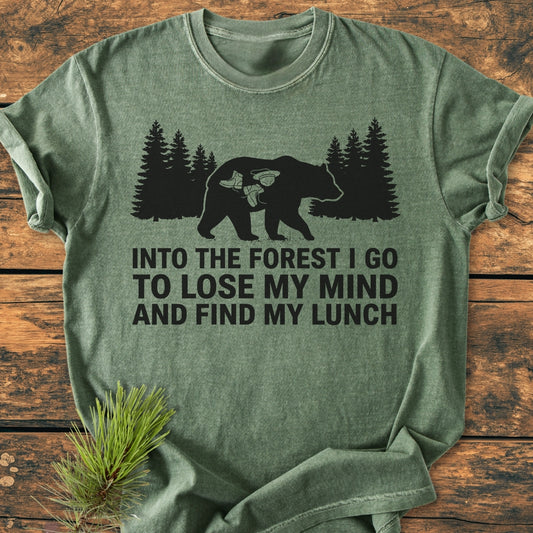 Into The Forest Bear - Vintage Wash Tee featuring a bear silhouette with hiking motifs and pine tree row, slogan about forest adventures; laid on rustic wood with pine sprig, highlighting its cozy, relaxed fit.