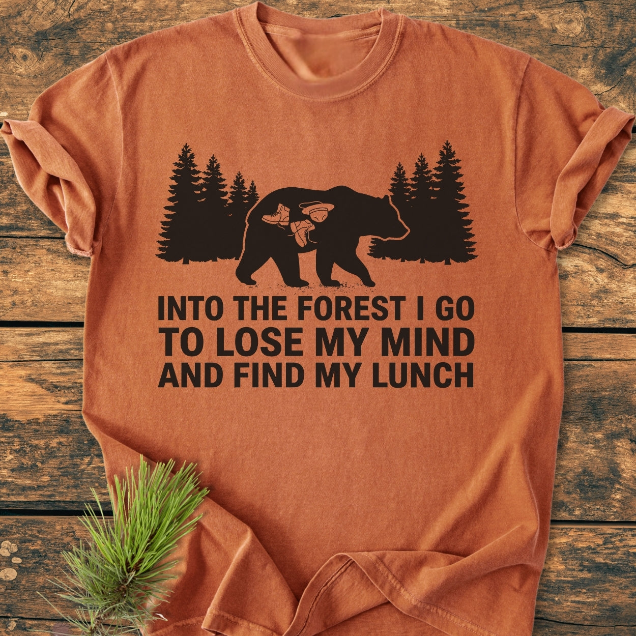 Into The Forest Bear - Vintage Wash Tee featuring a black bear graphic with hiking gear, and slogan, laid flat on a wooden surface with pine needles for an outdoor-inspired look.