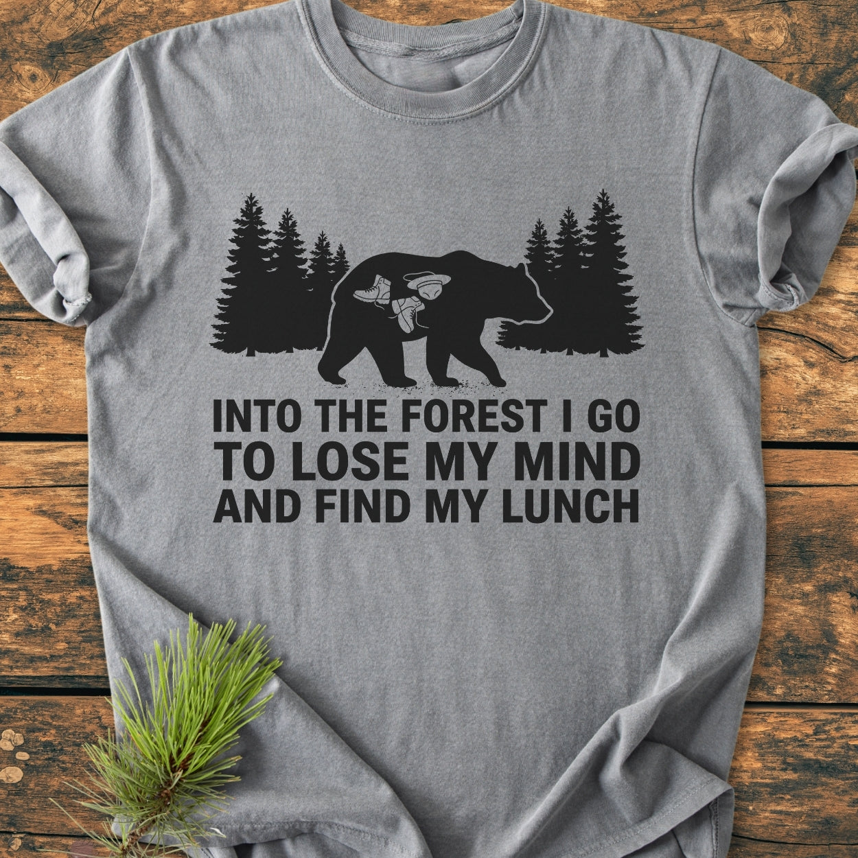 Heather-gray Into The Forest Bear - Vintage Wash Tee features a black bear silhouette with camping gear inside, set against evergreens, and a playful forest-themed slogan, styled on a wooden surface with pine.