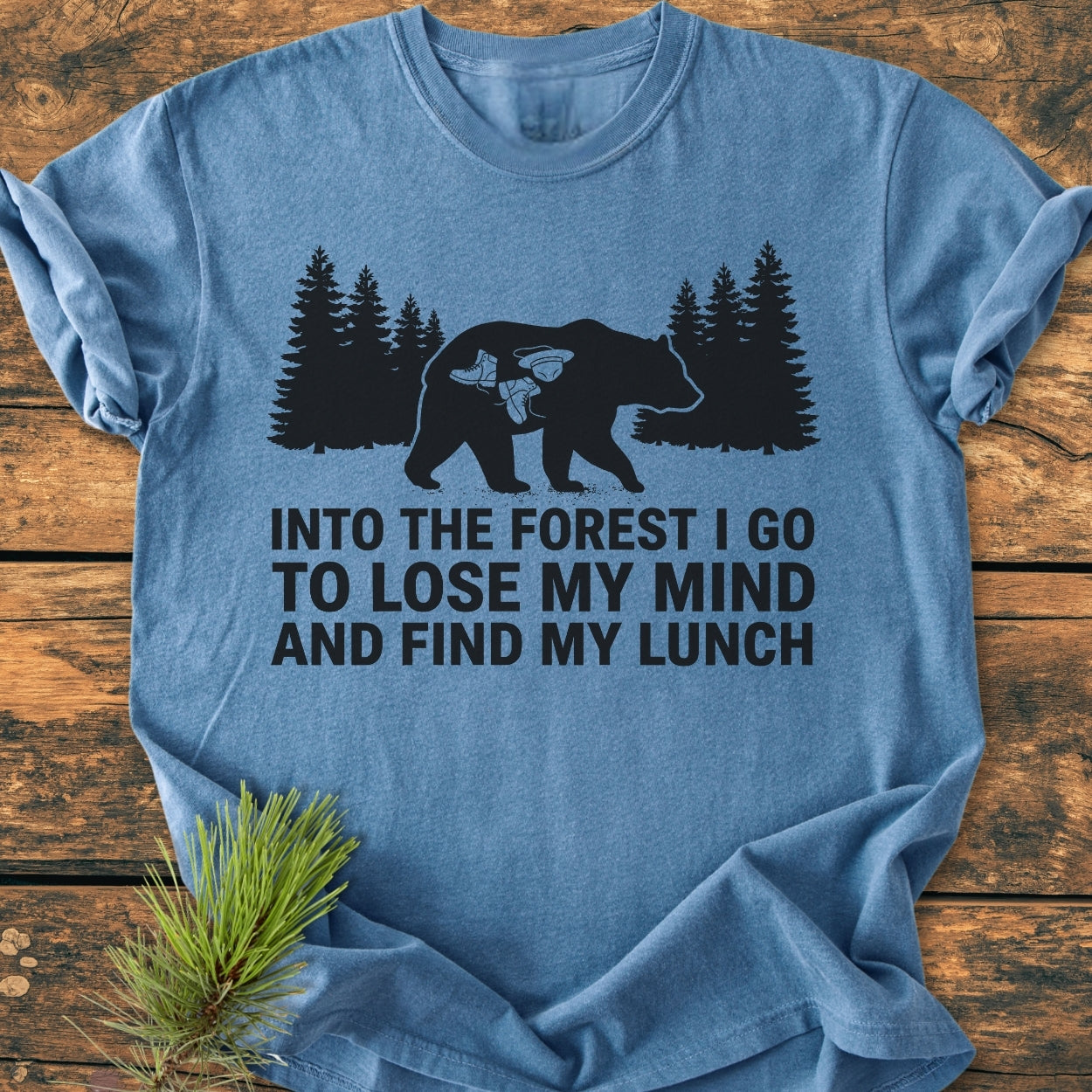 Into The Forest Bear - Vintage Wash Tee features a black bear graphic with hiking gear icons, overlaid with a nature-inspired slogan, displayed on a light-blue short-sleeve T-shirt on a wooden surface.