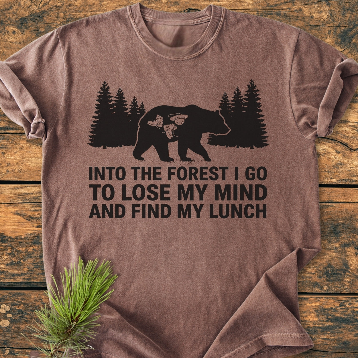Into The Forest Bear - Vintage Wash Tee features a bear silhouette with hiking motifs and a slogan, on a brown, short-sleeve T-shirt, styled with a pine branch on a wooden surface.