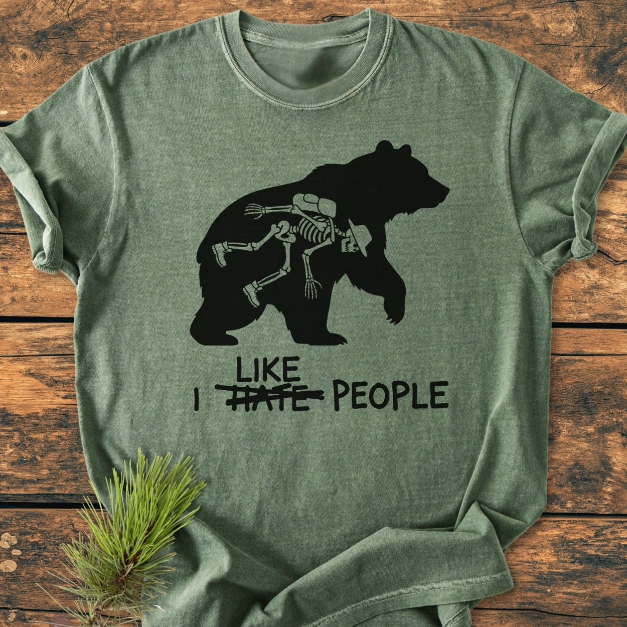 I Hate People - Vintage Wash Tee features a bear silhouette with a cartoon skeleton rider, bold text beneath, laid on wood, styled with a pine branch, showcasing a relaxed, soft-washed fit.