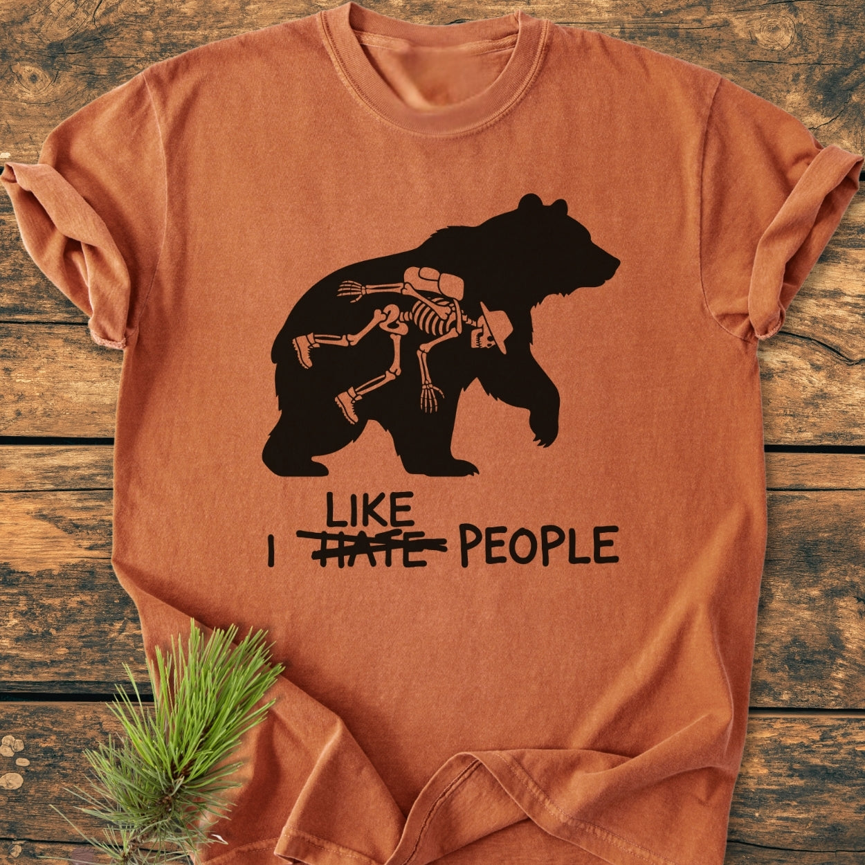 I Hate People - Vintage Wash Tee features a bear silhouette with a hiker skeleton inside. The phrase I ~HATE~ PEOPLE is beneath, styled with a pine sprig on a wooden surface.
