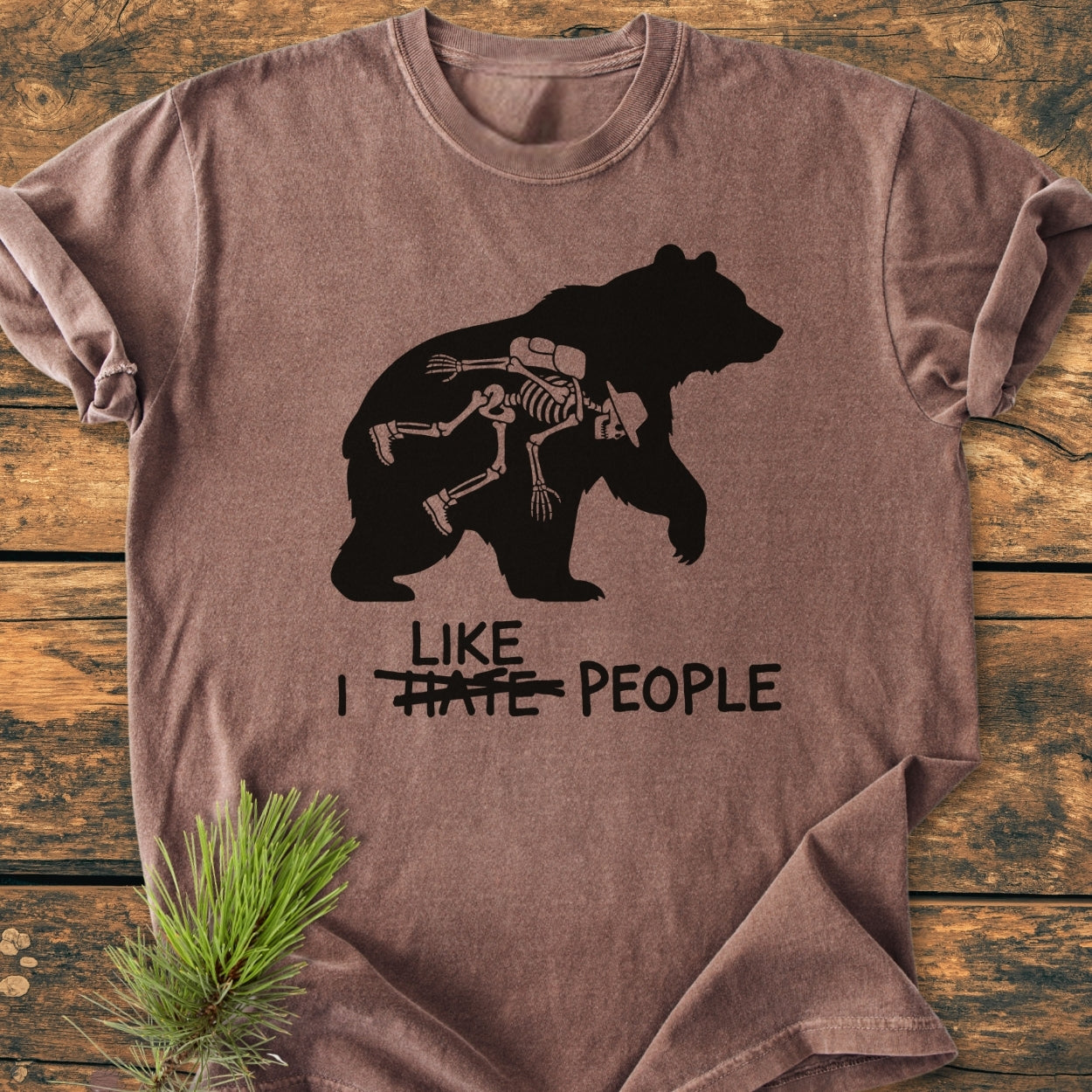 I Hate People - Vintage Wash Tee features a large black bear silhouette with a hiker skeleton inside. The phrase LIKE, I HATE PEOPLE has HATE crossed out, offering a playful design on a brown-washed T-shirt.