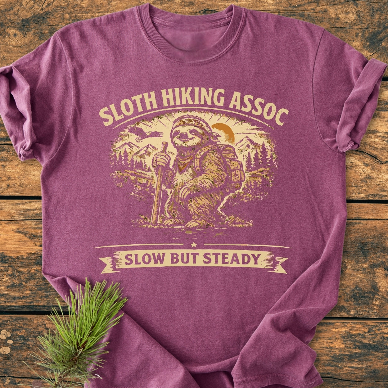 Sloth Hiking Assoc - Vintage Wash Tee features a sloth graphic with a bandana and backpack, hiking against mountains. This Comfort Colors T-shirt offers a relaxed fit and durable, soft-washed fabric.