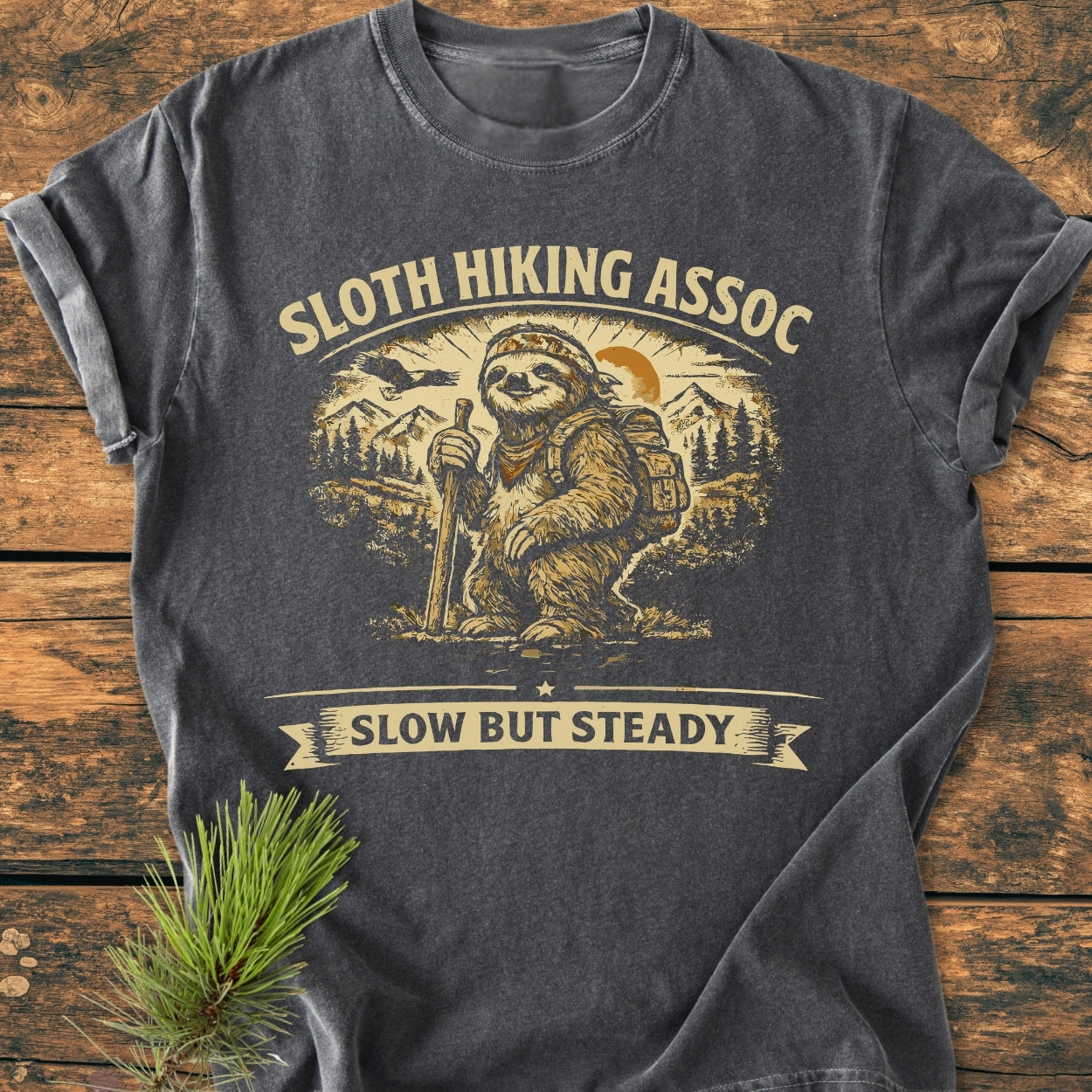 Sloth Hiking Assoc - Vintage Wash Tee features a cartoon sloth with a bandana and backpack against mountains and trees. The relaxed-fit, dark-gray shirt is laid on a wooden surface with a pine sprig beside it.
