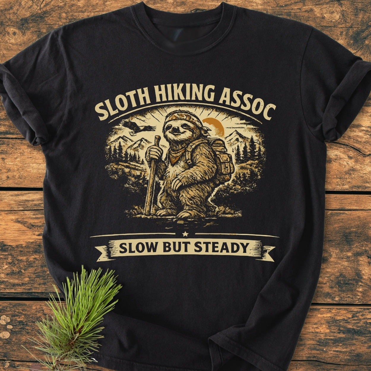 Sloth Hiking Assoc - Vintage Wash Tee featuring a sloth with bandana and backpack, hiking staff in hand, set against mountains and trees, with Slow But Steady banner, laid on wooden surface.