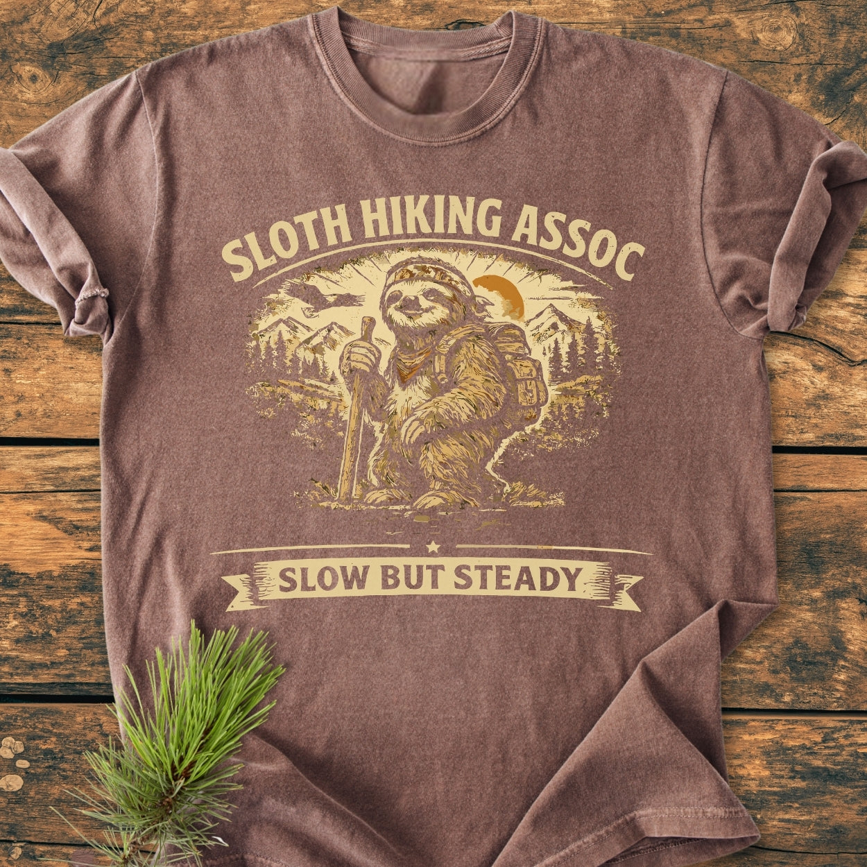 Sloth Hiking Assoc - Vintage Wash Tee features a hiking sloth graphic with text SLOTH HIKING ASSOC and SLOW BUT STEADY. Laid on wood, next to a pine sprig, highlighting its casual appeal.