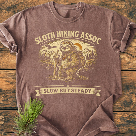 Sloth Hiking Assoc - Vintage Wash Tee features a hiking sloth graphic with text SLOTH HIKING ASSOC and SLOW BUT STEADY. Laid on wood, next to a pine sprig, highlighting its casual appeal.