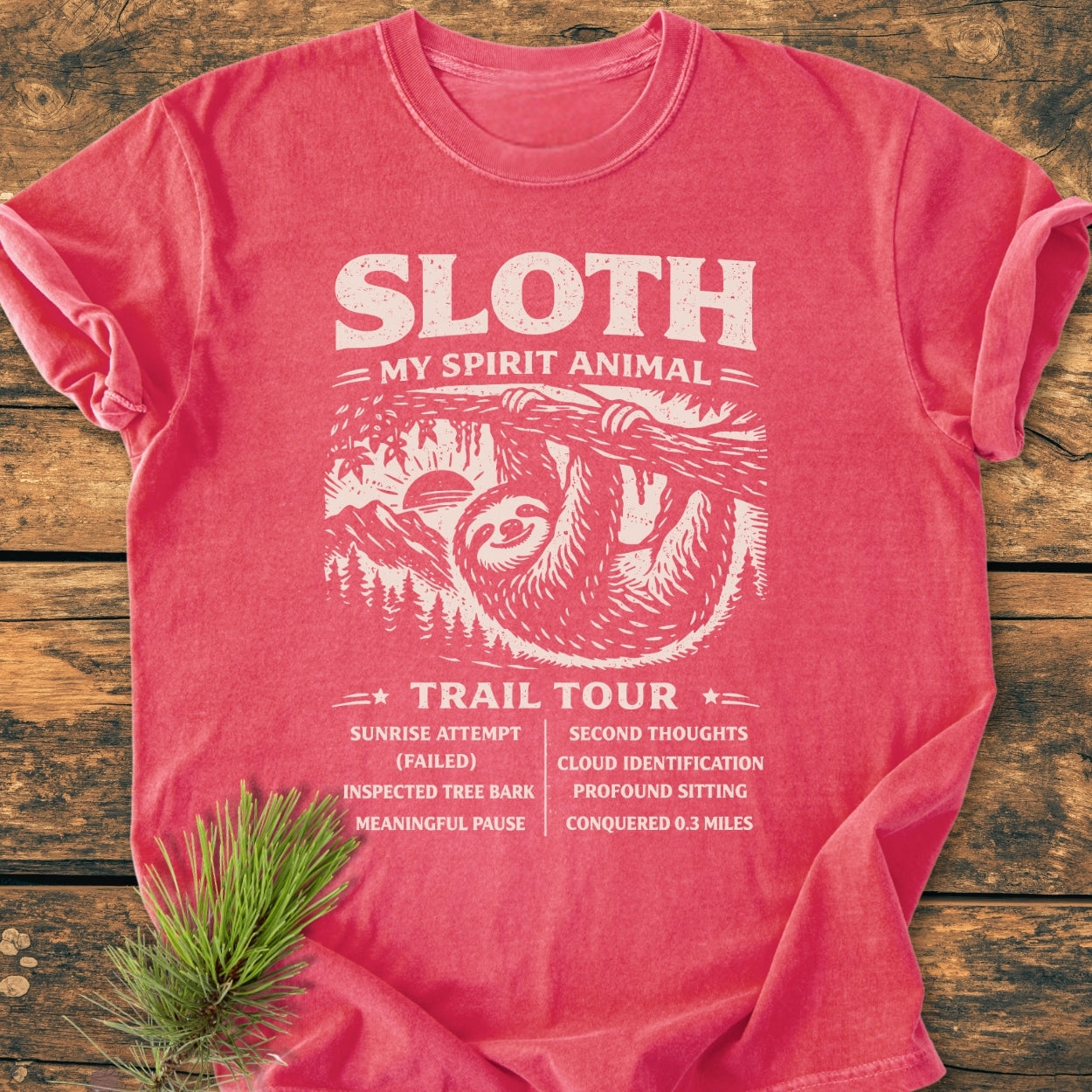 Sloth Spirit Animal Tour - Vintage Wash Tee featuring a whimsical sloth design with itinerary list; laid on wood, accented by pine branch, showcasing its relaxed fit and durable craftsmanship.